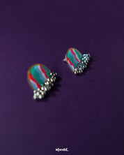 A pair of Aabshaar stud earrings displayed against a rich purple background, highlighting their semi-circle shape, hand-painted waves in teal, red, and white, and accented with oxidized silver ghungroos for a playful yet traditional touch — Muselot.