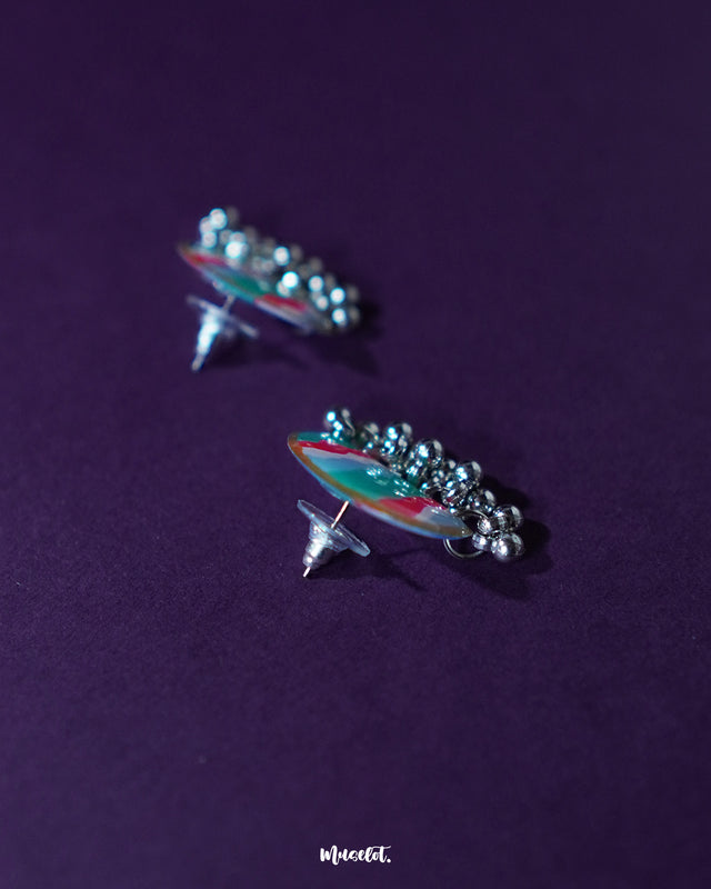 Flat-lay image of the Aabshaar stud earrings placed neatly on a dark purple surface, showcasing the vibrant hand-painted pattern in wavy teal, white, and red strokes, with antique silver ghungroo detailing adding a festive, folk-inspired finish — Muselot.
