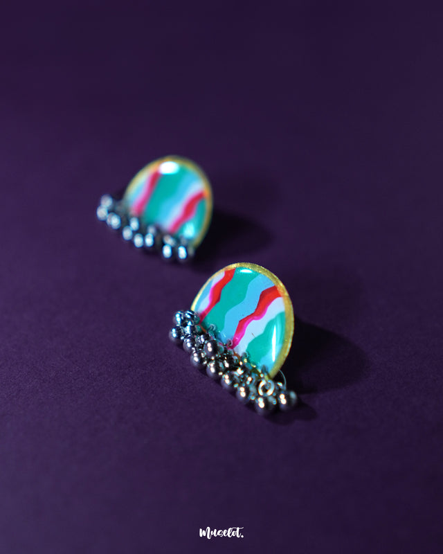 Aabshaar stud earrings positioned diagonally on a deep purple background, highlighting the fluid brushstrokes in teal, red, and white. The antique silver finish and tiny ghungroos add a graceful ethnic charm to this handmade design — Muselot.