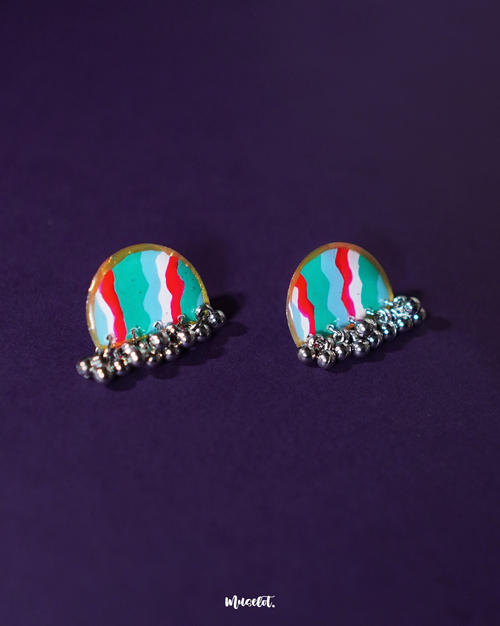 Aabshaar stud earrings displayed against a neutral backdrop, highlighting their floral pattern in vivid hues of teal, red, and white. The antique-finished base and tiny ghungroos below add a traditional charm to this intricate handmade pair — Muselot.