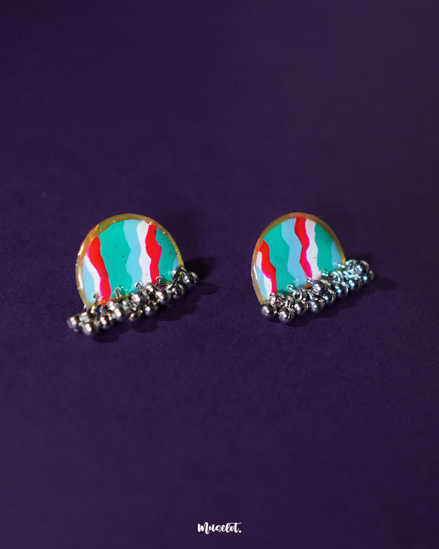 Aabshaar stud earrings displayed against a neutral backdrop, highlighting their floral pattern in vivid hues of teal, red, and white. The antique-finished base and tiny ghungroos below add a traditional charm to this intricate handmade pair — Muselot.