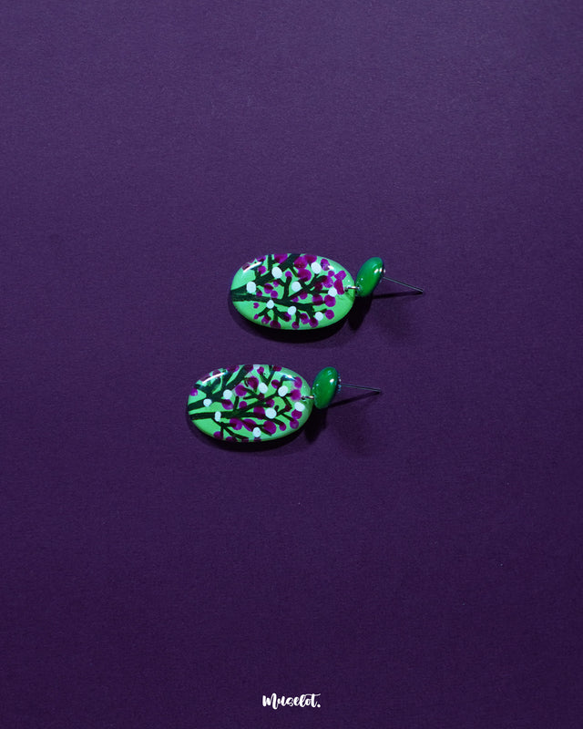 A pair of Baaghani green floral drop earrings displayed on a soft purple background, showcasing their vibrant green base and contrasting magenta-white floral detailing, highlighting the artisanal charm and playful elegance — Muselot.