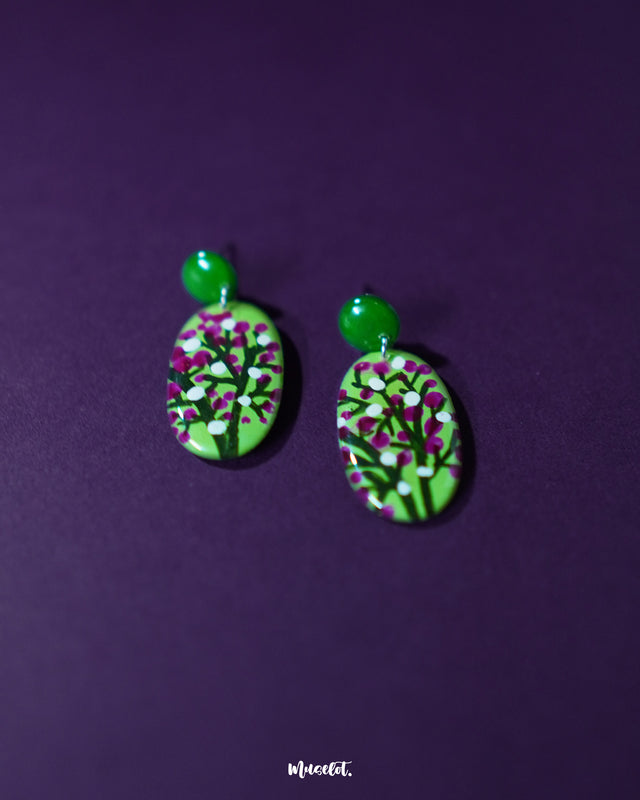 Close-up shot of Baaghani green floral drop earrings placed on a soft surface, highlighting the hand-painted floral accents in magenta and white over a green base, showcasing the intricate craftsmanship and vivid color contrast — Muselot.
