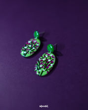 Baaghani green floral drop earrings placed against a purple background, showcasing their hand-painted white and magenta blossoms on a vibrant green base, highlighting the artistry and elegance — Muselot.