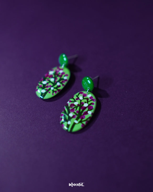 Baaghani green floral drop earrings placed against a purple background, showcasing their hand-painted white and magenta blossoms on a vibrant green base, highlighting the artistry and elegance — Muselot.