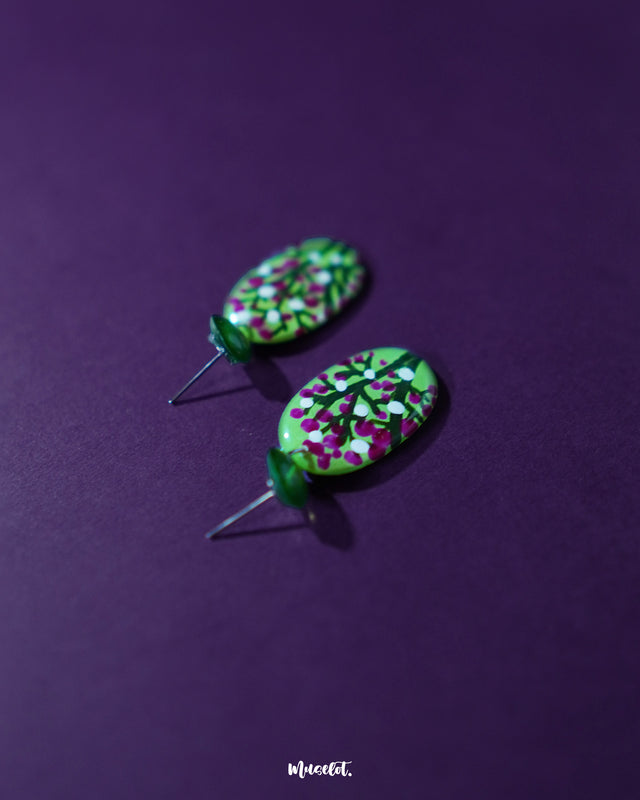 A pair of Baaghani green floral drop earrings displayed flat, featuring intricate hand-painted magenta and white blossoms with dark stems, showcasing vibrant contrast and artisanal finesse — Muselot.