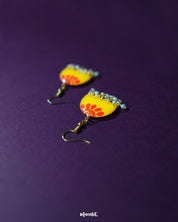 Close-up of Basantika yellow floral hook earrings placed on a deep purple background, highlighting the intricate red petal pattern and delicate light blue bead drops for a playful ethnic accent — Muselot.