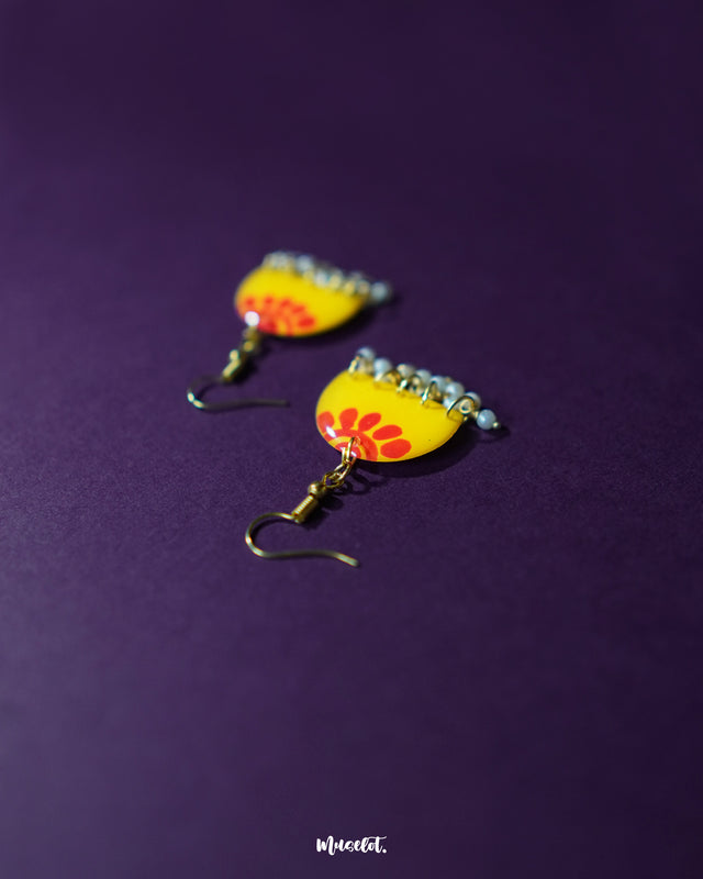 Close-up of Basantika yellow floral hook earrings placed on a deep purple background, highlighting the intricate red petal pattern and delicate light blue bead drops for a playful ethnic accent — Muselot.