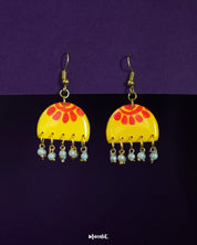 A pair of Basantika yellow and red hook earrings against a purple background, showcasing the semicircle design with bold red motifs and delicate dangling blue beads for a festive, handcrafted charm — Muselot.