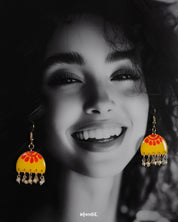 A woman wears the Basantika yellow and red hook earrings, highlighting the hand-painted semicircle motif in warm hues and delicate blue bead danglers that add a touch of artisanal flair — Muselot.