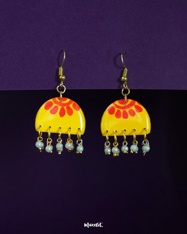 A pair of Basantika yellow and red hook earrings against a purple background, showcasing the semicircle design with bold red motifs and delicate dangling blue beads for a festive, handcrafted charm — Muselot.