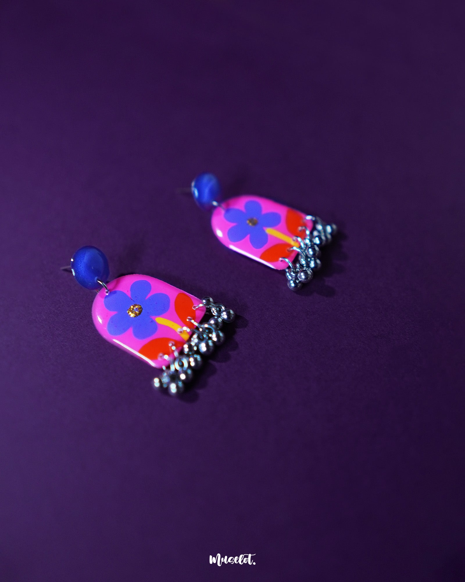 Benazir floral earrings with hand-painted pink and red petals, delicately crafted in a vibrant drop design, set against a rich purple background that highlights their bright, cheerful tones and intricate detailing — Muselot