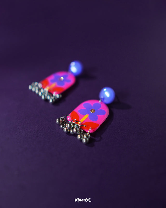 Benazir floral earrings placed on a soft purple background, featuring purple flowers with deep red leaves and a glossy finish that highlights their handmade charm — Muselot.
