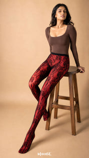 The model layers black rose designer lace stockings over red opaque stockings while seated, showcasing the bold red-and-black floral contrast of the Sinful Rose combination – Muselot.