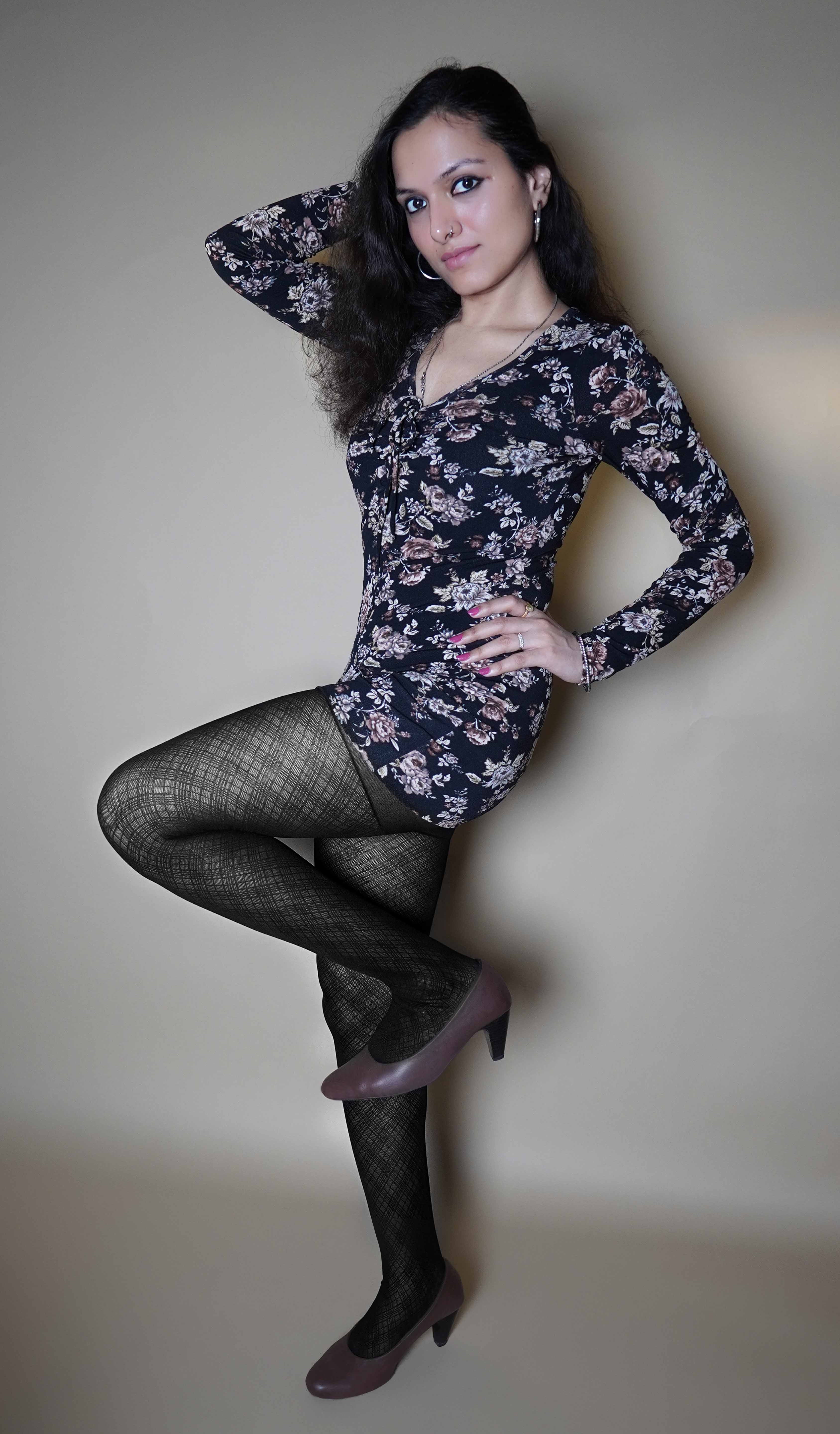 A woman wearing patterned black colored tights for women, posing with her hand on her waist and a leg bent upwards as she stands sideways, looking at the camera, highlighting the stretchable durable material-Muselot