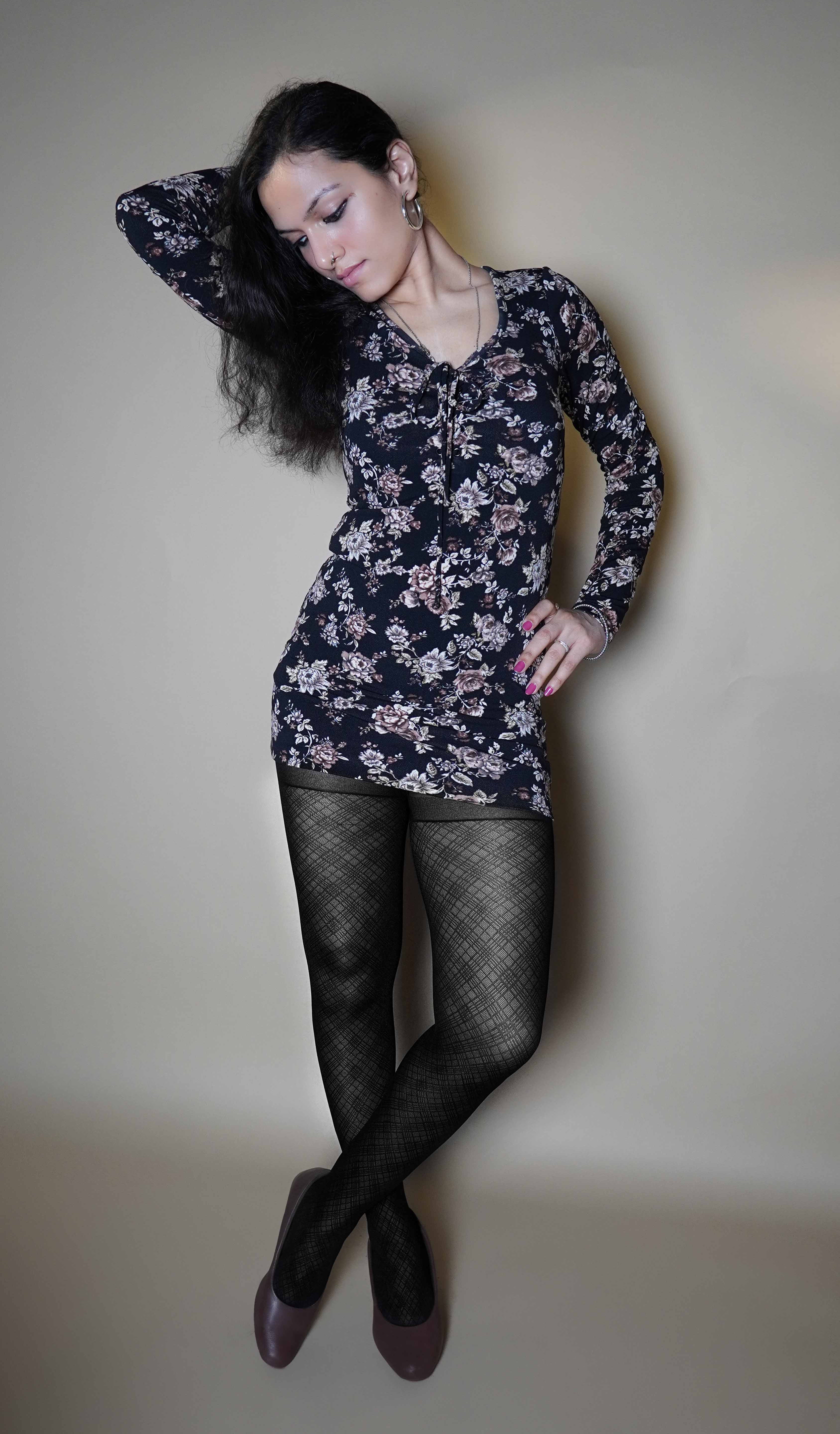 A woman wearing black transparent sheer patterned stockings, posing with her hand on her waist and other behind her head as it is bent sideways, standing cross legged, highlighting the relaxed modern look-Muselot