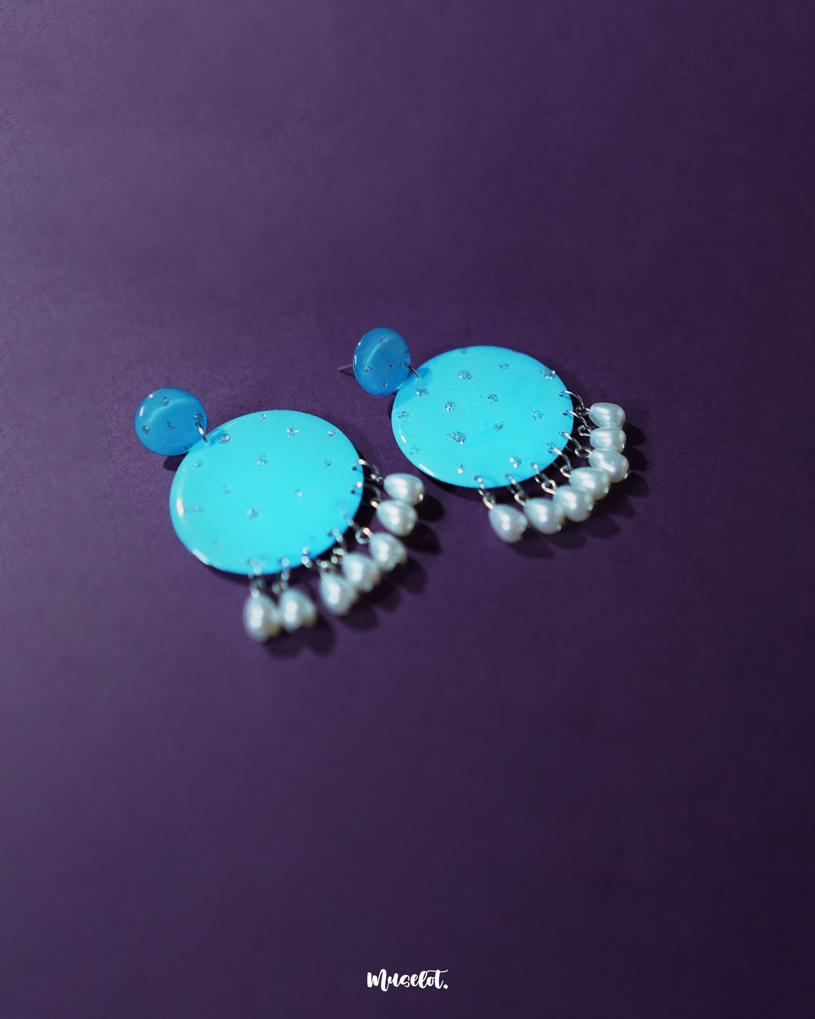 Blue pearl earrings displayed on a purple background, featuring small and large round turquoise blue discs with subtle silver glitter accents, finished with dangling white pearl-like beads – Muselot.