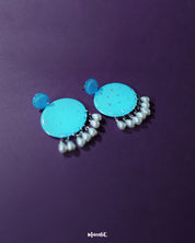 Blue pearl earrings displayed on a purple background, featuring small and large round turquoise blue discs with subtle silver glitter accents, finished with dangling white pearl-like beads – Muselot.