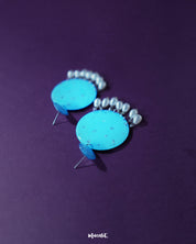 Blue pearl earrings featuring small and large turquoise blue discs with subtle silver glitter accents, complemented by dangling white pearl-like beads, artistically showcased to emphasize their vibrant color and elegant detailing – Muselot.