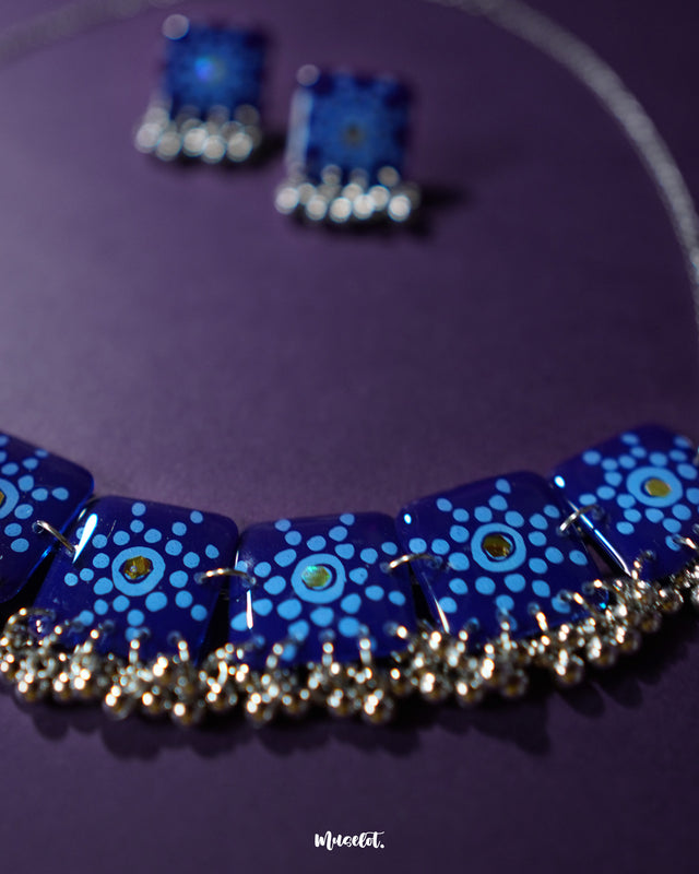 Falakzaar blue necklace and earring set displayed against a purple background, showcasing the hand-painted detailing and highlighting the elegant craftsmanship and vibrant charm of this statement set — Muselot.