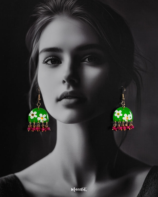 A model wearing green floral hook earrings featuring hand-painted white blossoms on a glossy green base, accented with magenta bead danglers—highlighting cheerful charm and showcasing handcrafted finesse — Muselot.