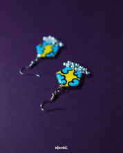 Front view of Gulaftab hook earrings against a purple background, showcasing the vibrant blue floral design on a yellow base with dangling silver beads for a playful yet artistic charm — Muselot.