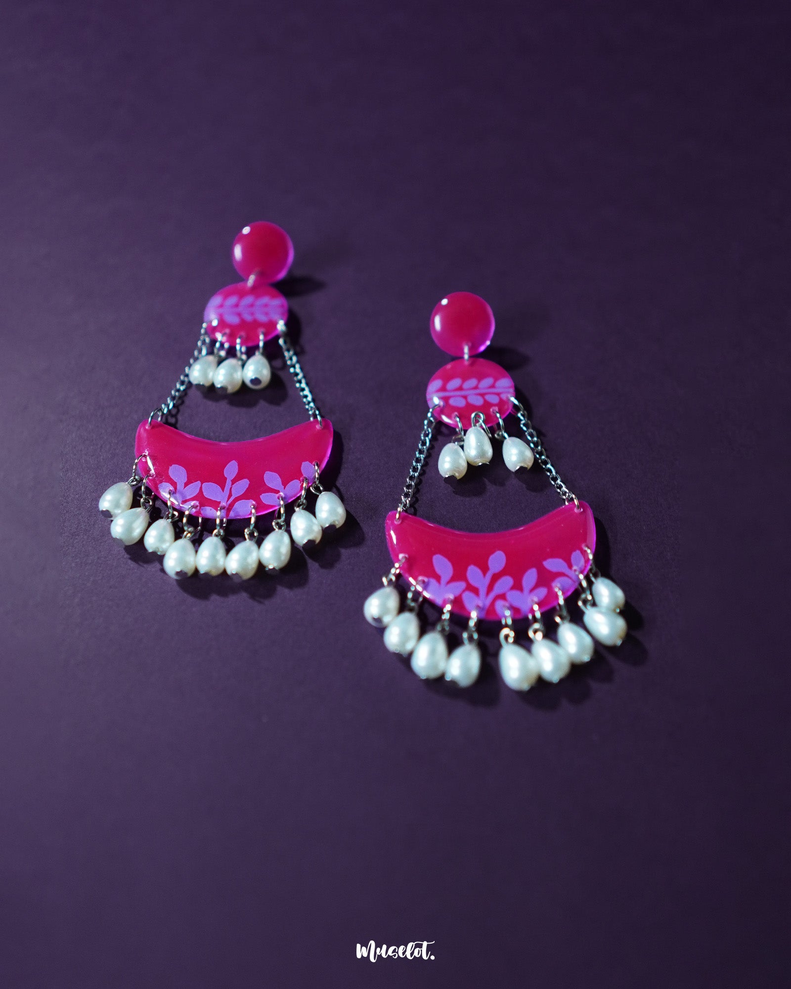 A pair of Gulfaam pink pearl chandbali earrings displayed against a deep purple background, featuring glossy magenta tiers with lilac leaf motifs and cascading pearl drops that add a festive sparkle — Muselot.
