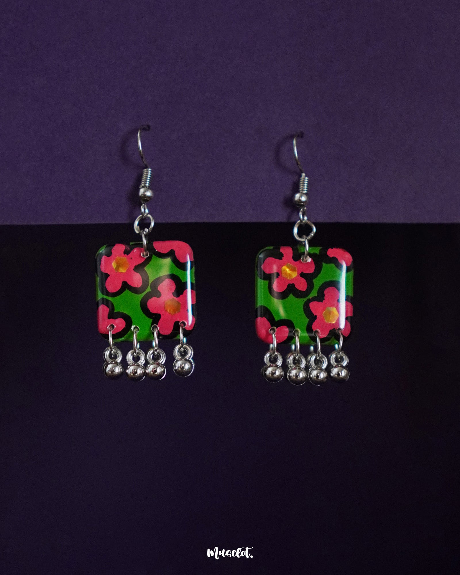 Gulnaar floral earrings featuring rectangular charms with vibrant pink and red flowers on a green background, outlined in black, and adorned with delicate silver bead tassels, beautifully displayed against a deep purple backdrop – Muselot.
