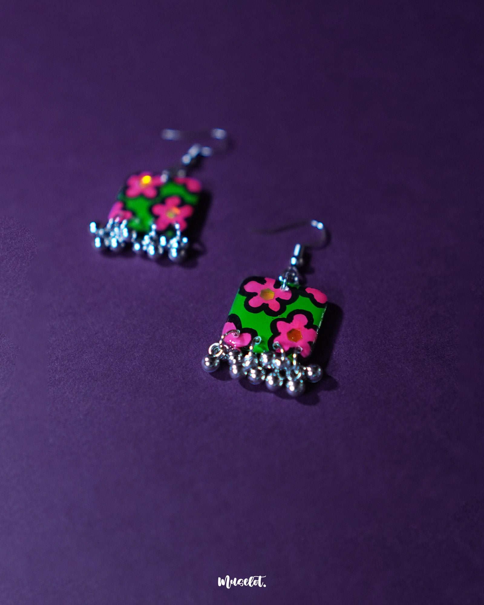 Gulnaar handmade floral hook earrings displayed on a purple background, featuring rectangular charms with vibrant pink and red flowers on a green background outlined in black, accented with delicate silver bead tassels – Muselot.