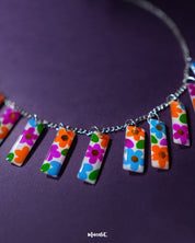 A closer view of the Gulraas floral necklace set captures the colorful hand-painted panels in bright hues of orange, pink, blue, and green, all suspended on a sleek silver chain—highlighting the playful charm and artisanal elegance of this handcrafted accessory — Muselot.