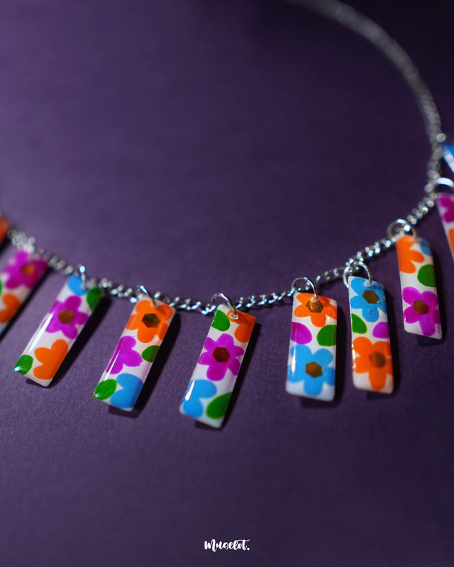 A closer view of the Gulraas floral necklace set captures the colorful hand-painted panels in bright hues of orange, pink, blue, and green, all suspended on a sleek silver chain—highlighting the playful charm and artisanal elegance of this handcrafted accessory — Muselot.