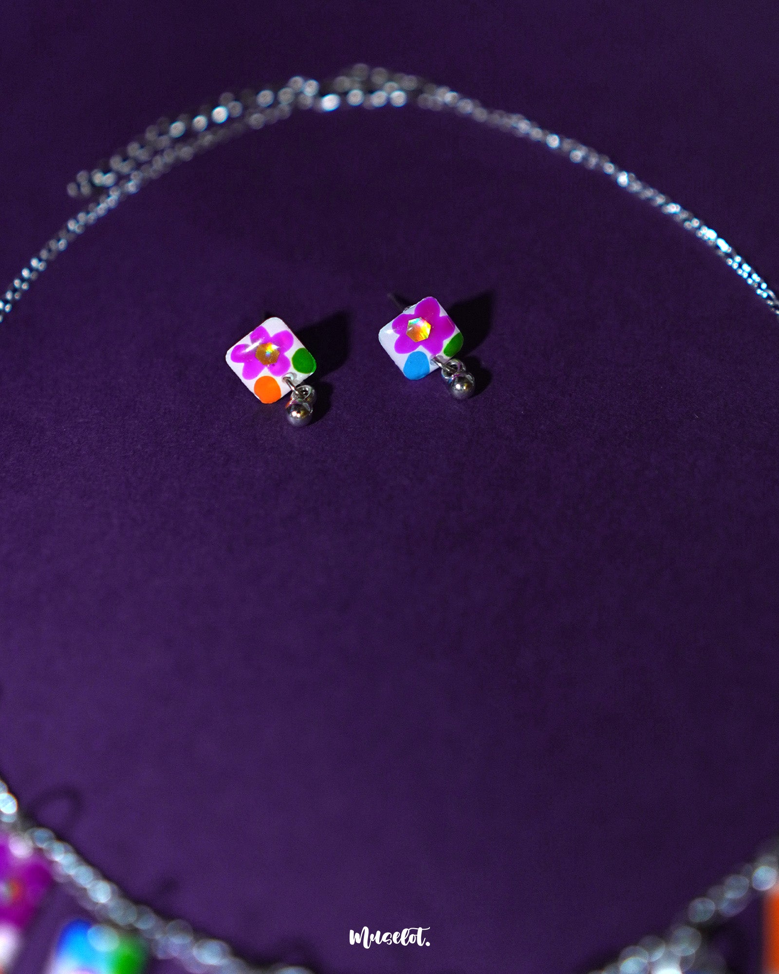 A pair of floral stud earrings from the Gulraas necklace set is displayed against a deep purple background, highlighting their vibrant hand-painted petals in shades of pink, yellow, and violet—radiating playful elegance in a compact form — Muselot.