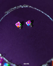 A pair of floral stud earrings from the Gulraas necklace set is displayed against a deep purple background, highlighting their vibrant hand-painted petals in shades of pink, yellow, and violet—radiating playful elegance in a compact form — Muselot.