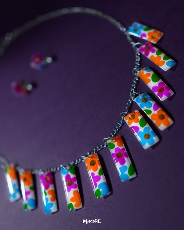 A top-view of the Gulraas floral necklace set is displayed on a purple backdrop, highlighting the vivid rectangular floral tiles in shades of orange, blue, purple, and green, paired with matching stud earrings for a coordinated, cheerful ensemble — Muselot.