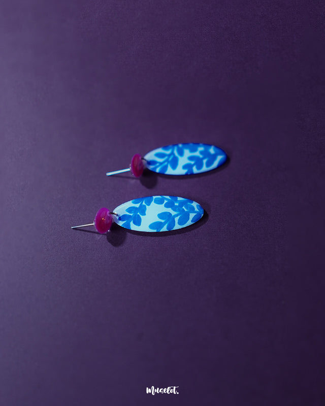 Gulsaba blue handmade drop earrings displayed on a purple background, featuring glossy red circular studs paired with elongated white oval drops hand-painted with bold blue leafy patterns – Muselot.