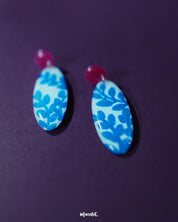 Close-up of the Gulsaba blue handmade drop earrings, showcasing glossy red circular studs paired with elongated white oval drops detailed with bold hand-painted blue leafy patterns, highlighting the intricate brushwork and vibrant contrast – Muselot.
