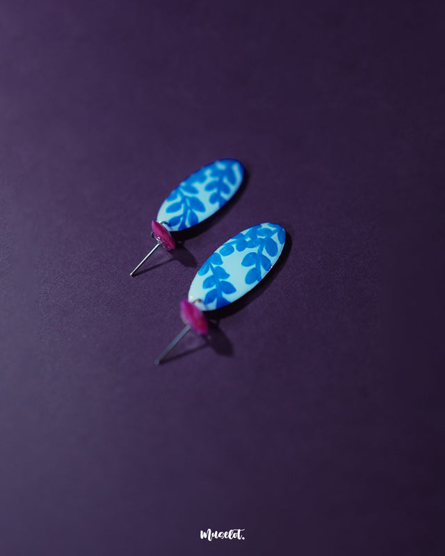 Top view of the Gulsaba blue handmade drop earrings, featuring glossy red circular studs attached to elongated white oval drops adorned with bold hand-painted blue leafy patterns, capturing their artistic detailing and vibrant color contrast – Muselot.