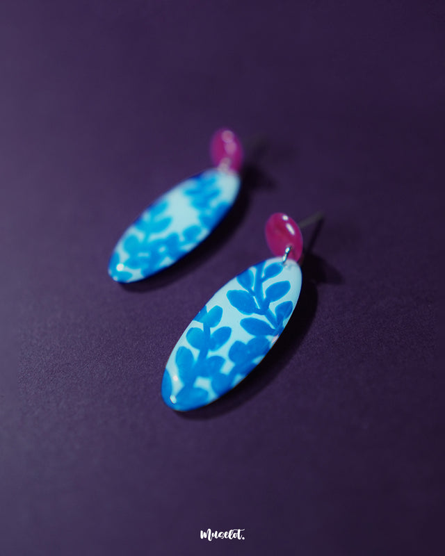 Side close-up view of the Gulsaba blue handmade drop earrings, showing glossy red circular studs paired with elongated white oval drops decorated with bold hand-painted blue leafy patterns, highlighting their detailed craftsmanship and striking color contrast – Muselot.