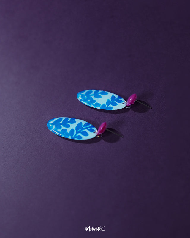 Gulsaba blue handmade drop earrings featuring glossy red circular studs with elongated white oval drops adorned with bold hand-painted blue leafy patterns, artistically displayed to showcase their vibrant, contemporary design – Muselot.