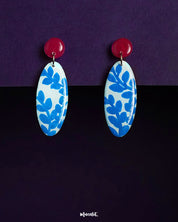 Gulsaba drop earrings featuring glossy red circular studs paired with elongated oval drops in white, adorned with hand-painted blue leafy patterns, beautifully displayed against a deep purple background – Muselot.
