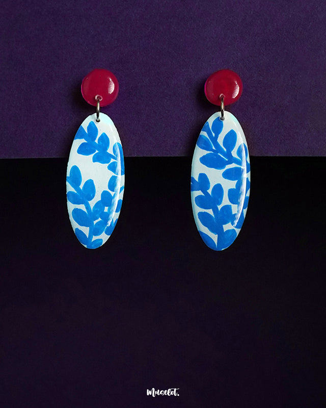 Gulsaba drop earrings featuring glossy red circular studs paired with elongated oval drops in white, adorned with hand-painted blue leafy patterns, beautifully displayed against a deep purple background – Muselot.