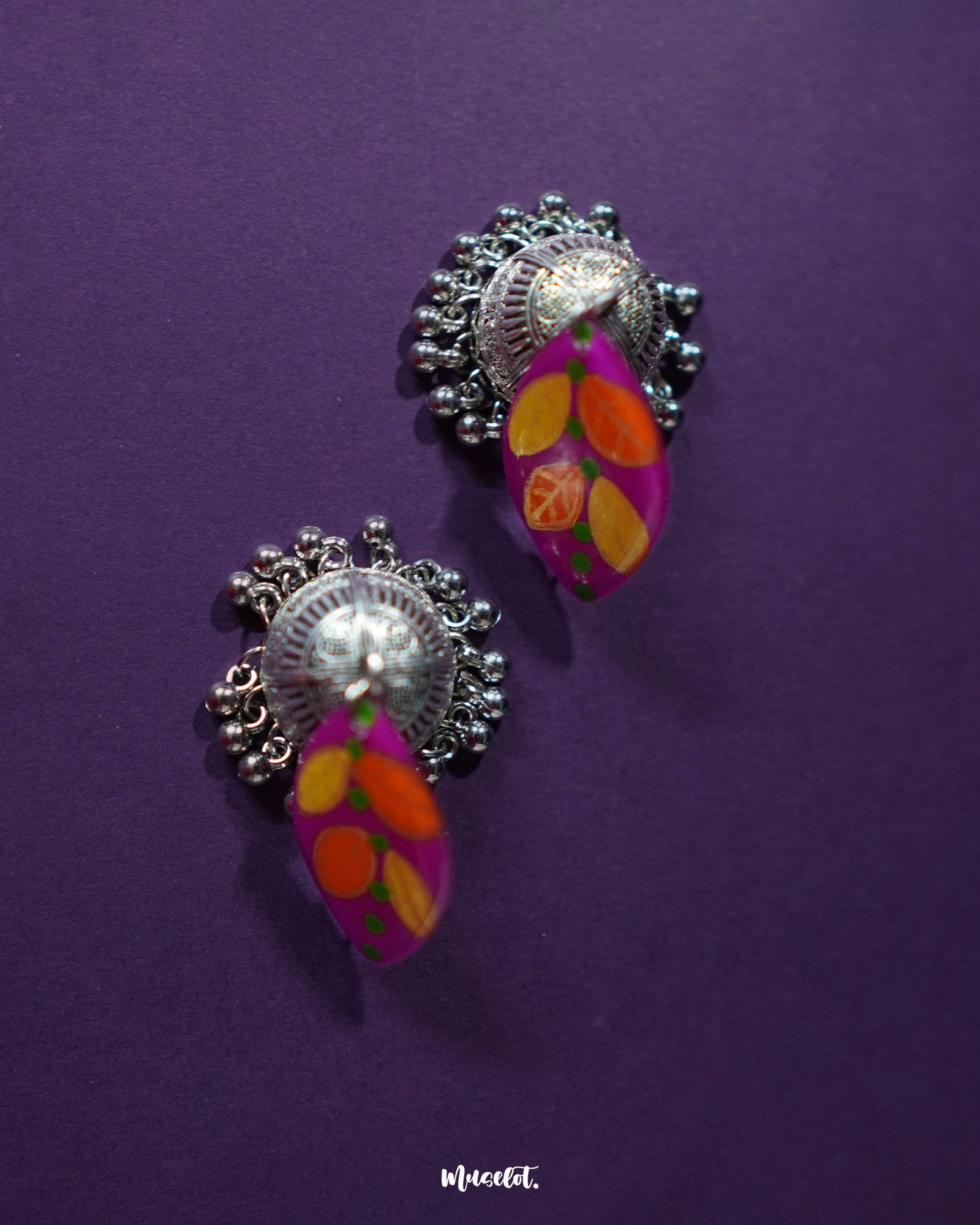 Gulzarish floral jhumkas displayed on a purple background, featuring marquise-shaped tops painted in vibrant purple with orange and yellow leaf motifs accented by green dots, paired with intricately carved oxidized silver dome-shaped drops and delicate hanging beads – Muselot.