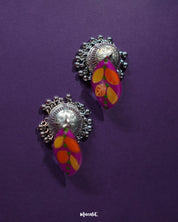 Gulzarish floral jhumkas displayed on a purple background, featuring marquise-shaped tops painted in vibrant purple with orange and yellow leaf motifs accented by green dots, paired with intricately carved oxidized silver dome-shaped drops and delicate hanging beads – Muselot.