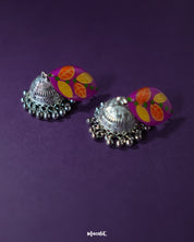 Gulzarish floral jhumkas featuring marquise-shaped purple tops with orange and yellow leaf motifs accented by green dots, paired with intricately detailed oxidized silver dome-shaped drops and delicate hanging beads, beautifully showcased to highlight their vibrant and traditional design – Muselot.