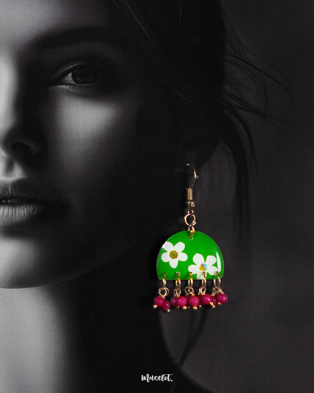 A woman wears handmade hook earrings with a vibrant green base and hand-painted white flowers, finished with magenta bead danglers—showcasing playful elegance and highlighting artisanal craftsmanship — Muselot