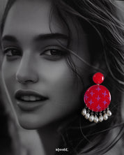 A woman wearing handmade stud drop earrings in vibrant pink, designed with circular patterns and dangling pearl-like drops, showcasing artistic craftsmanship and highlighting statement style — Muselot.