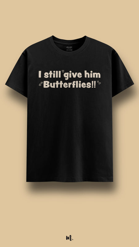 Black graphic t-shirt showcasing the statement “I still give him Butterflies!!” in bold beige typography with small heart details, highlighting a sweet, romantic, and playful tone against a neutral beige background – Muselot