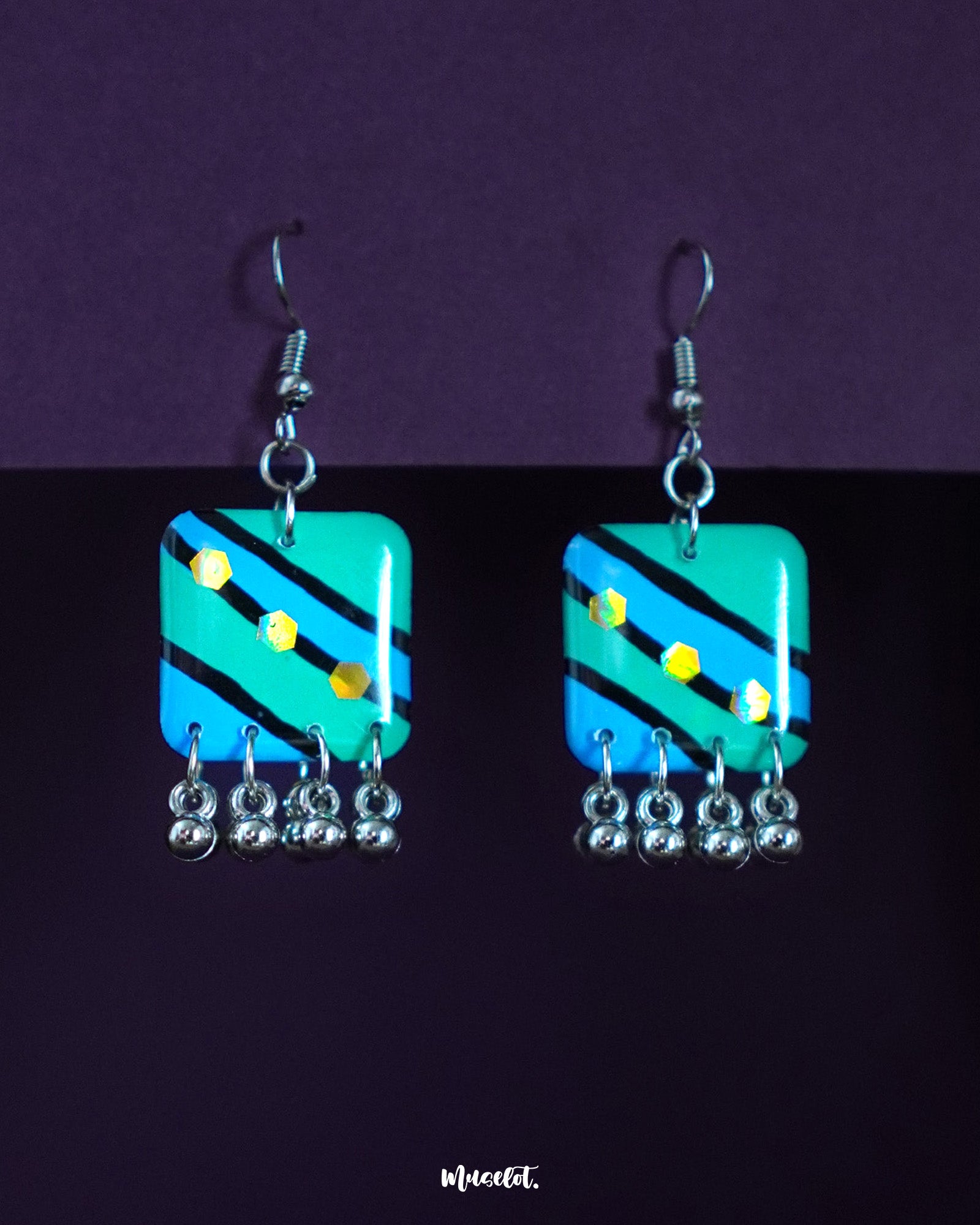 A pair of Jaltarang strip drop earrings featuring hand-painted in vivid green and blue diagonal stripes, adorned with yellow dot motifs and finished with silver-toned ghungroos that add a soft jingle—Muselot.