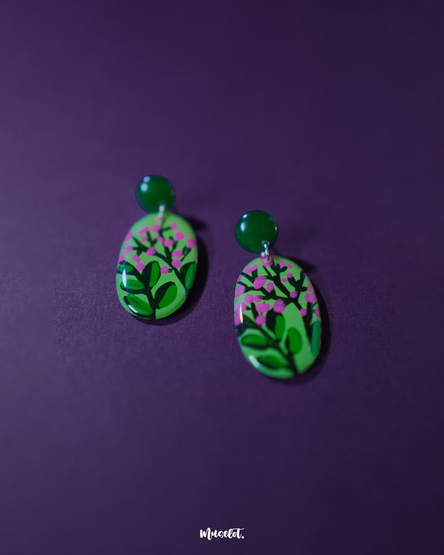 A pair of Kalibaan drop earrings laid flat on a soft neutral backdrop, highlighting the bold green and pink floral artwork. The handcrafted detailing and vivid colors showcase a perfect blend of boho elegance and playful charm — Muselot.
