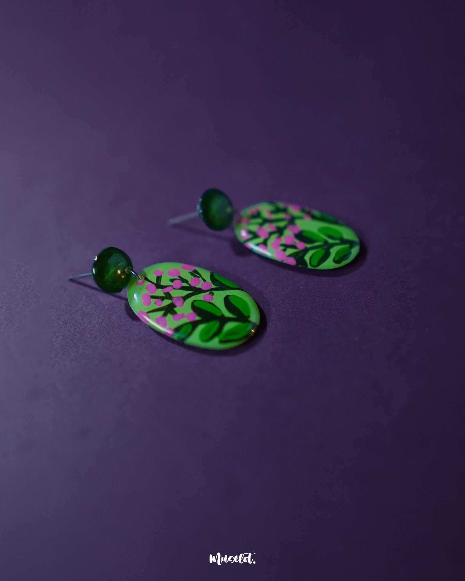 A close-up view of the Kalibaan drop earrings resting on a textured surface, highlighting the vibrant hand-painted floral design in shades of green and pink. The detailing showcases artisanal craftsmanship and colorful statement appeal — Muselot.